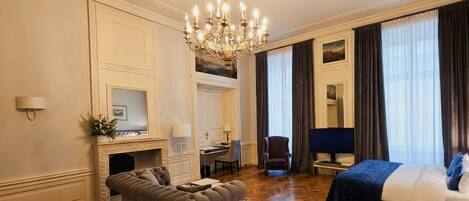 A hotel room with a fireplace, a sofa, a bed, a desk, and a chandelier.