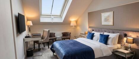 A hotel room with a bed, a desk, two chairs, a small table, a nightstand, a TV, and a skylight.