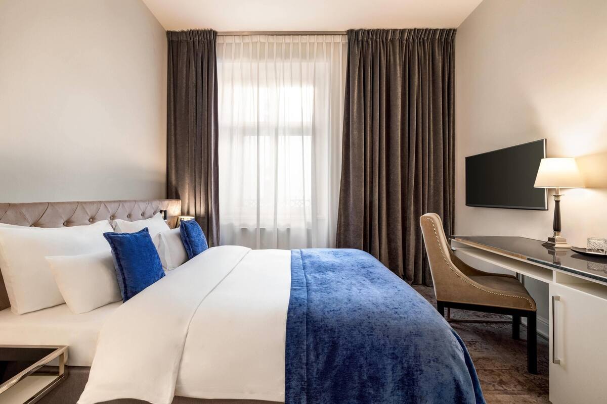 Hotellihuone, jossa on sänky, työpöytä, tuoli, televisio ja ikkuna, jossa on verhot.