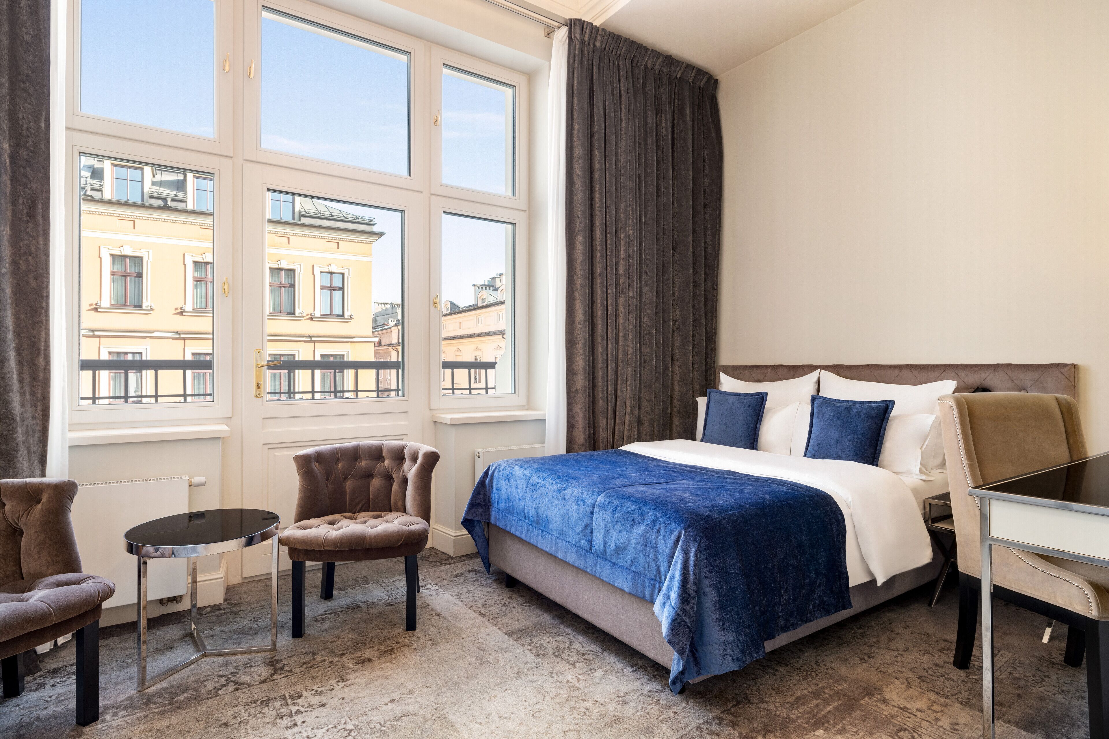 Une chambre d’hôtel avec un grand lit, une petite table ronde, deux fauteuils et une vue sur les immeubles à travers de grandes fenêtres.