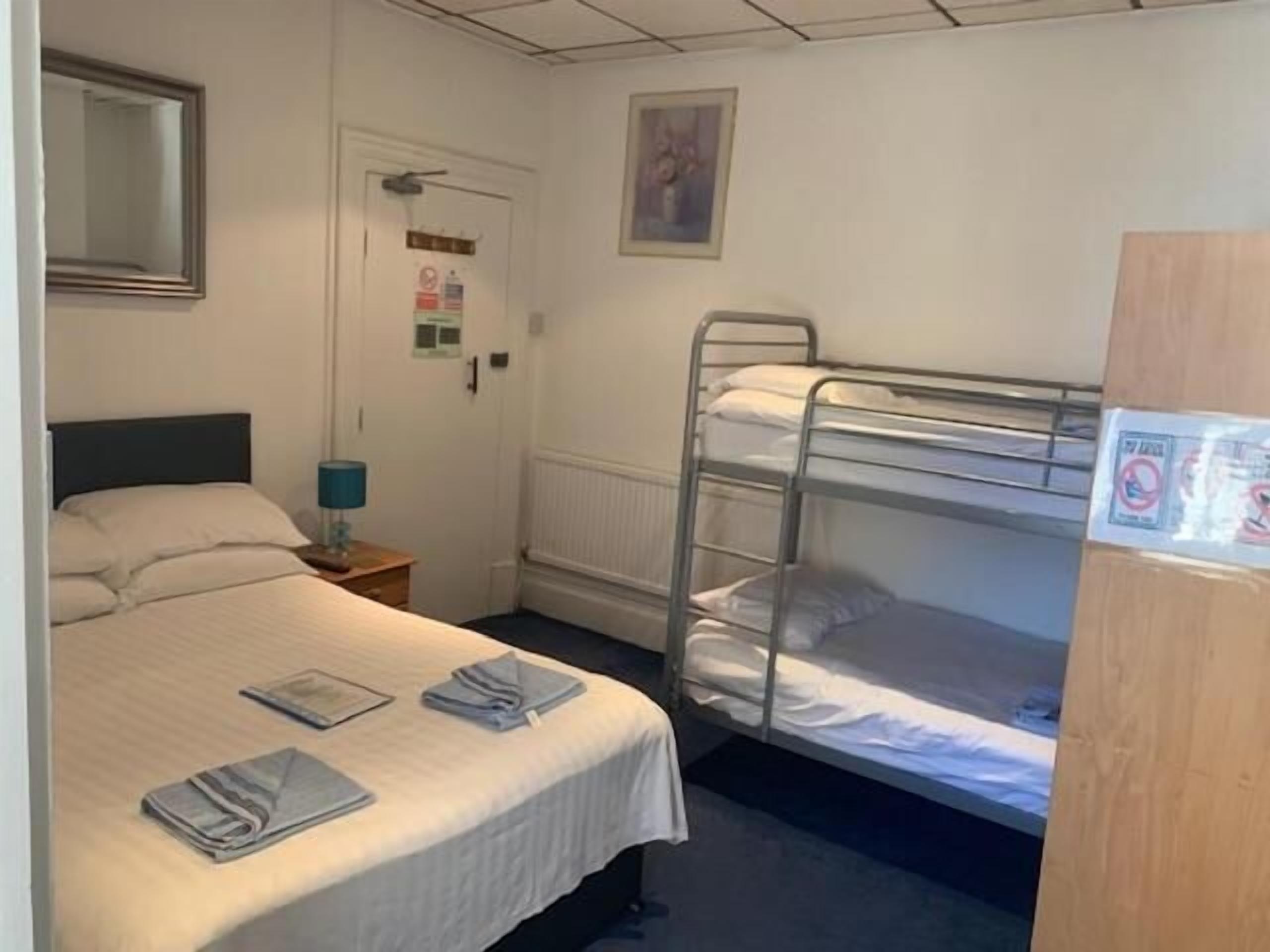 Un lit simple avec une couette blanche et deux oreillers, une table de chevet avec une lampe, un lit superposé avec du linge de lit blanc, une porte avec une pancarte et un tableau encadré au mur.