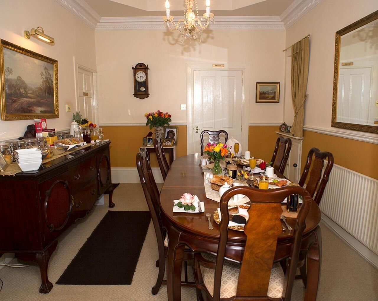 A dining room with a long wooden table, chairs, a chandelier, a clock, and a painting on the wall.