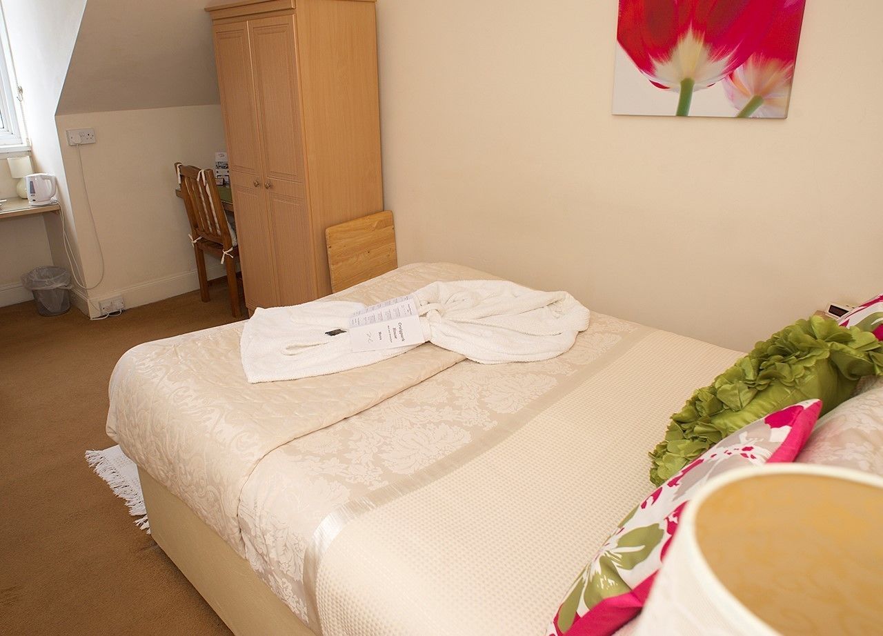 A neatly made bed with a white duvet and a folded towel, a wooden wardrobe, a chair, and a painting of a tulip on the wall.