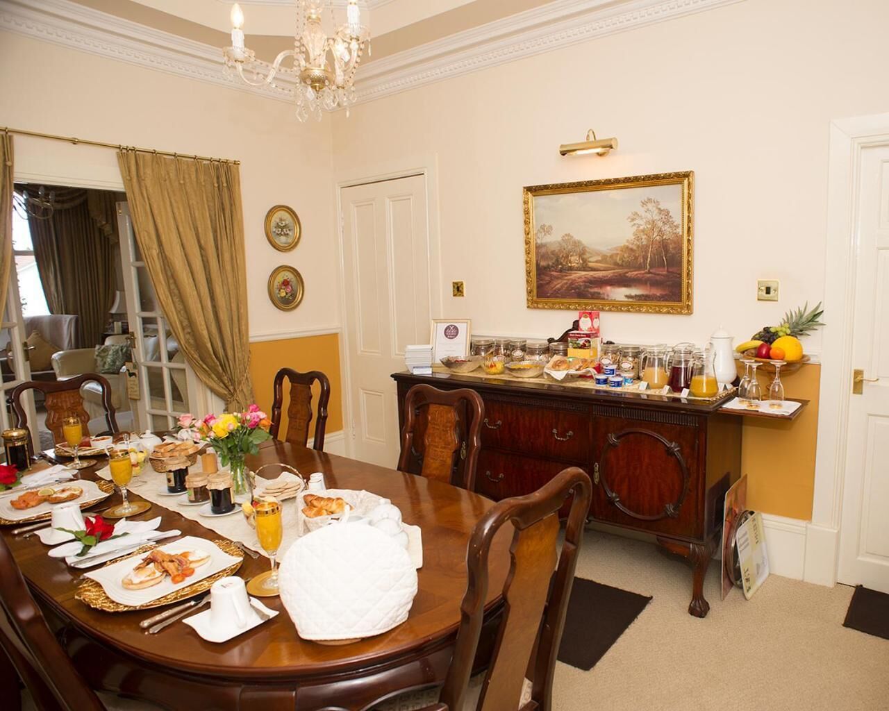 A dining room with a wooden table set for breakfast, a buffet with various food items, and a painting on the wall.