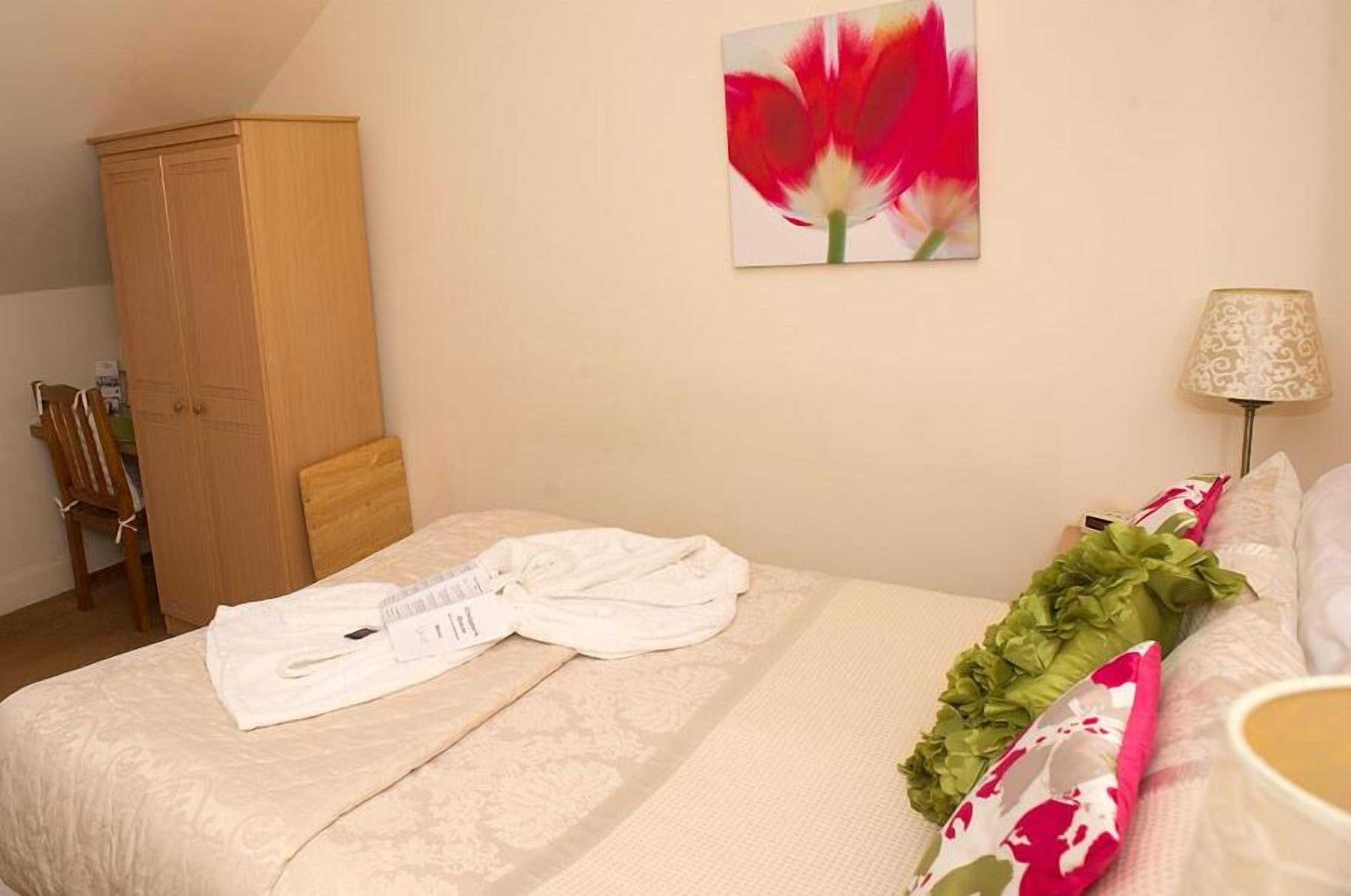 A hotel room with a bed, a wooden wardrobe, a painting of a flower, a lamp, and a green and pink pillow.