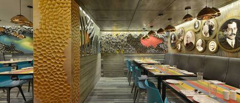 A restaurant with a long communal table, blue chairs, and a wall decorated with framed portraits.