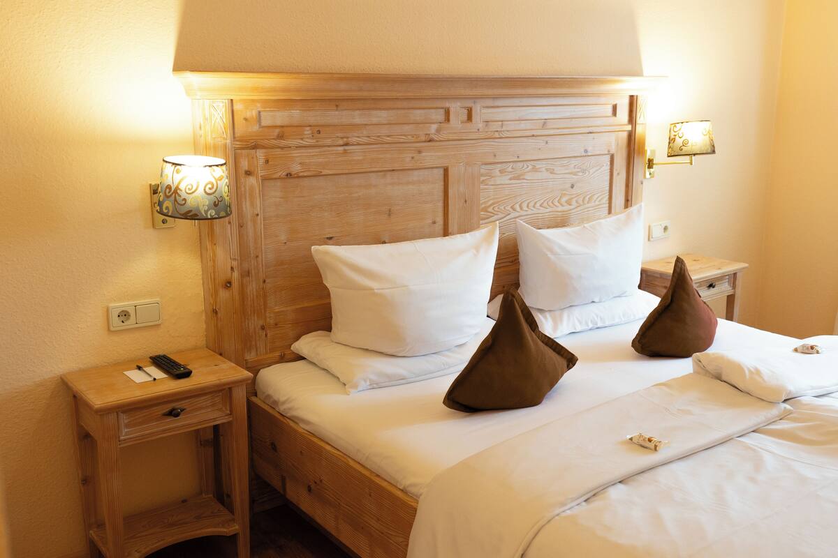 Un lit bien fait, avec des draps blancs, deux oreillers et une télécommande posée sur une table de chevet en bois.