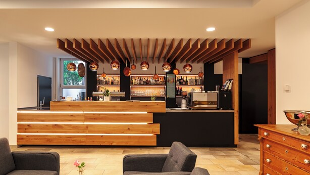 Un espace bar moderne agrémenté d’éléments en bois, un coin salon avec des canapés et un comptoir de bar où sont exposées diverses bouteilles et différents types de verres.