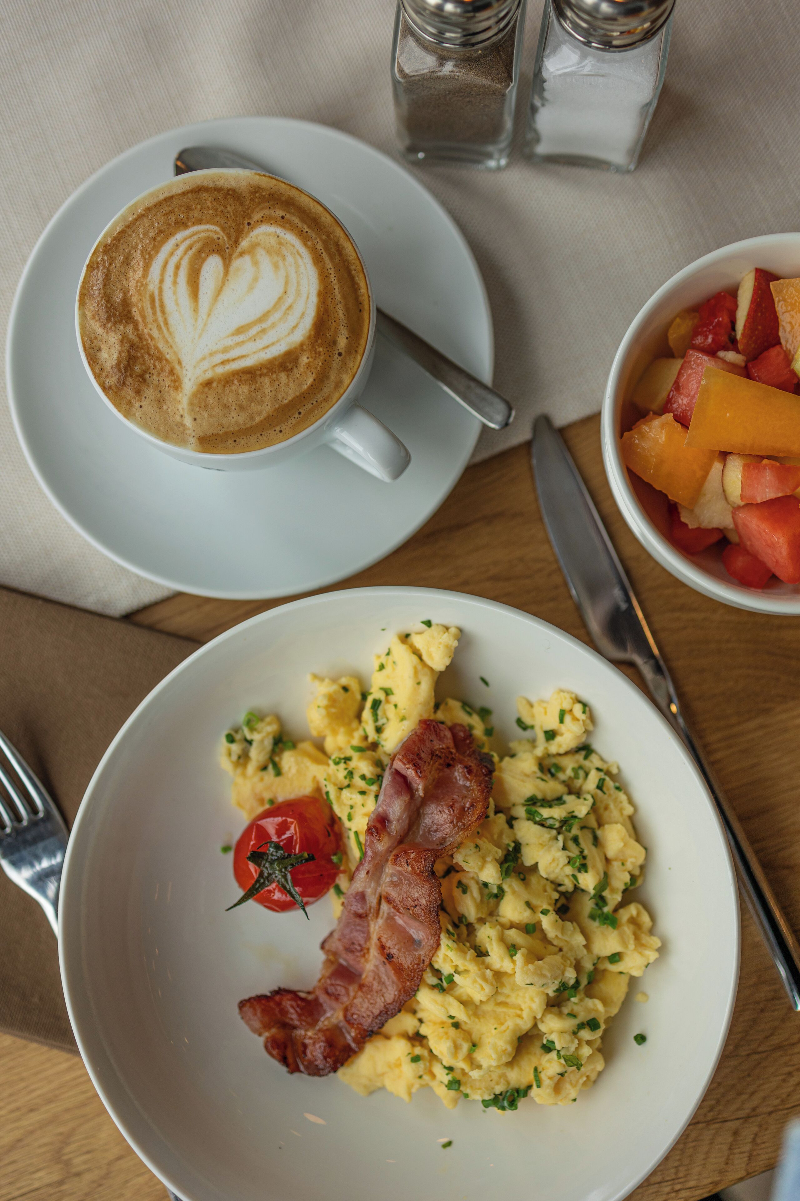 A breakfast setting with scrambled eggs, bacon, a tomato, and a cup of coffee with latte art.