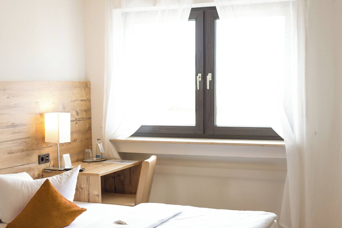 Une chambre d’hôtel avec un lit, une table de chevet, une lampe, une fenêtre avec des rideaux et une tête de lit en bois.
