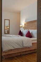 Una cama bien hecha con sábanas blancas y dos almohadas rojas, un cabecero de madera, un espejo fijado a la pared y una lámpara de noche.