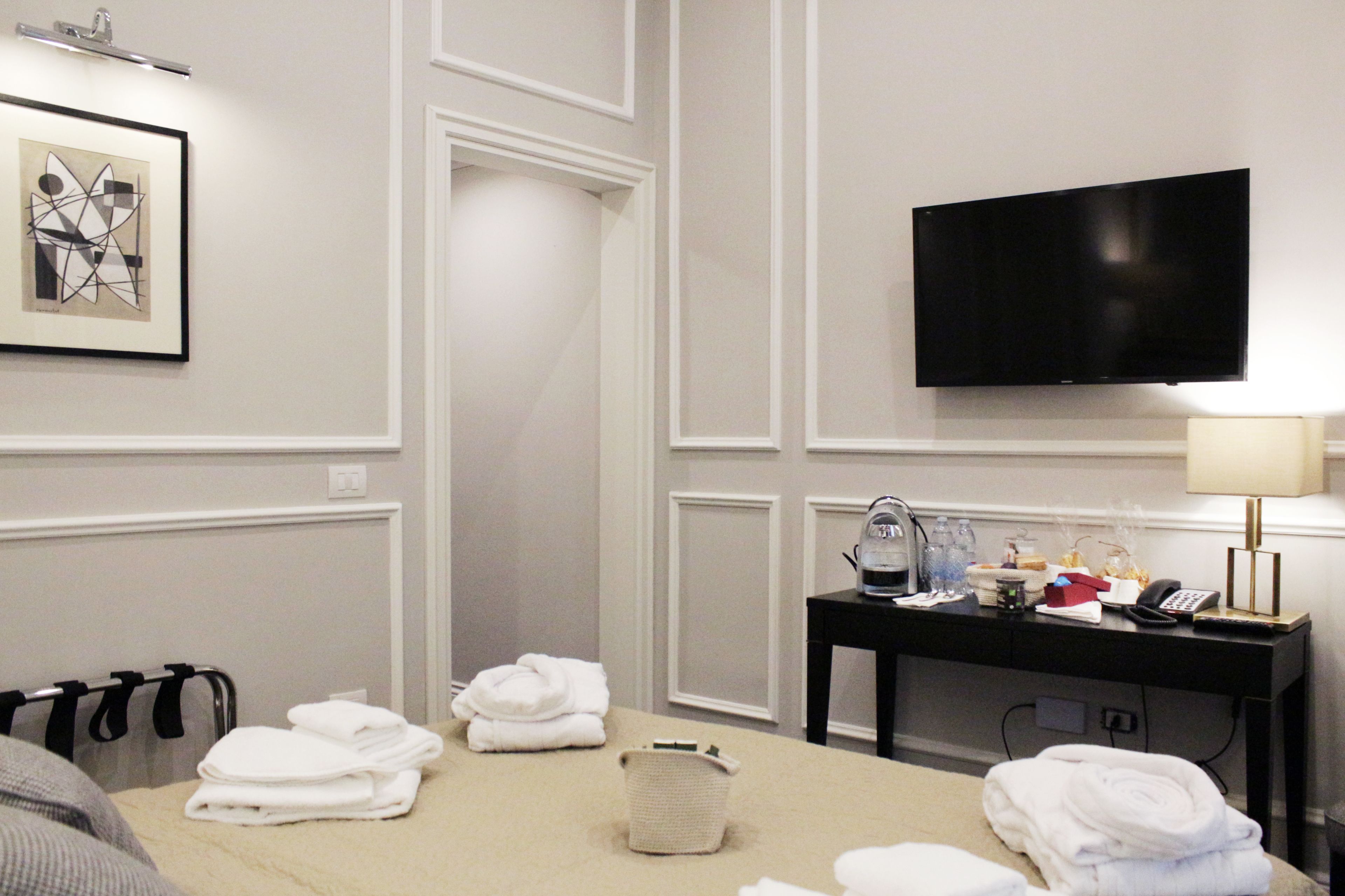 A hotel room with a bed, a desk with a lamp, a flat-screen TV, and a framed artwork on the wall.