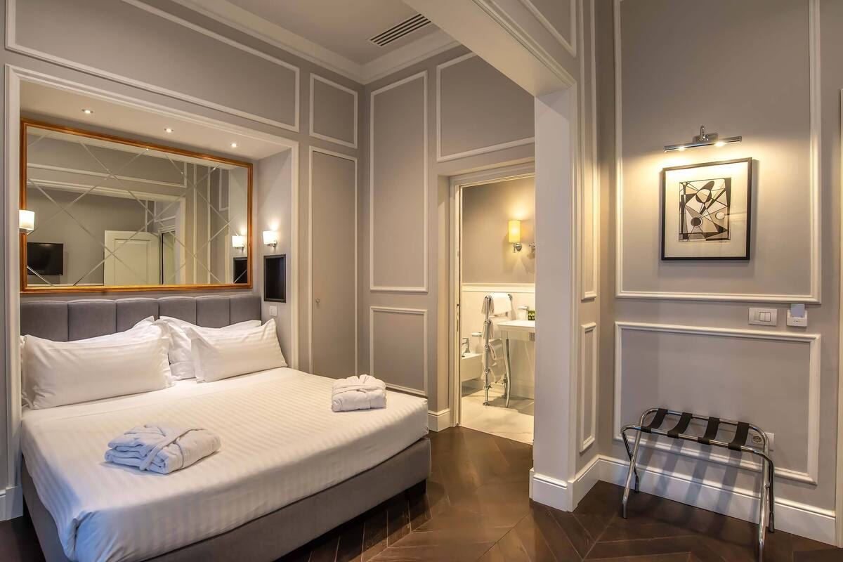 A hotel room with a large bed, a mirror, a bedside table, a bench, and a bathroom visible through an open door.