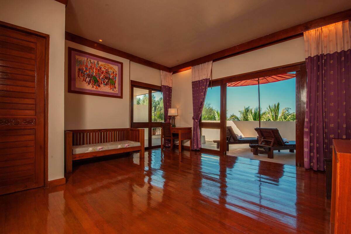 A spacious room with a wooden floor, a bench, a table, and a chair. There is a large window with curtains and a view of palm trees.
