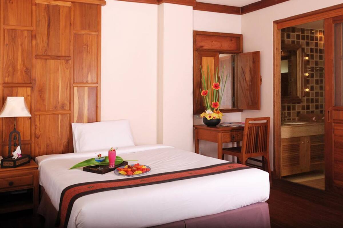A hotel room with a large bed, a wooden nightstand with a lamp, a desk with a chair, and a bathroom visible through an open door.
