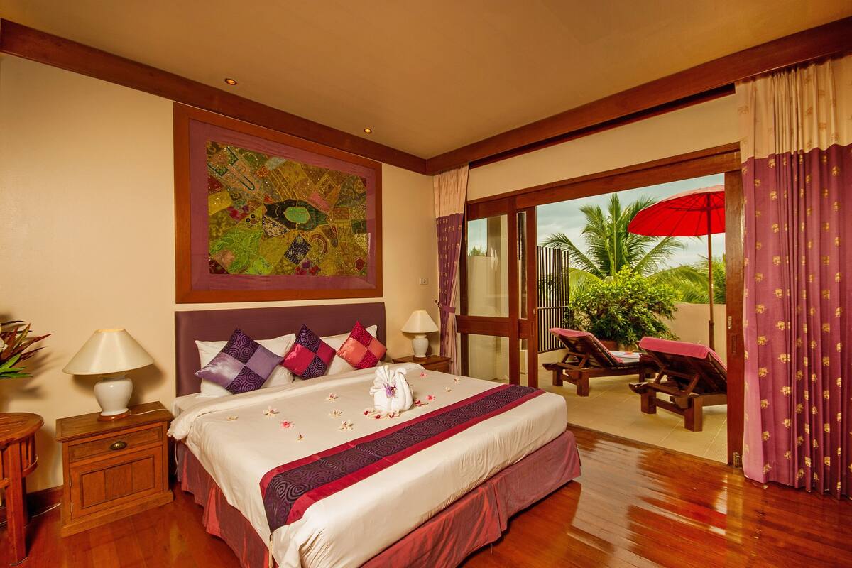 A hotel room with a large bed, two bedside tables, a nightstand, a window with curtains, a red umbrella outside, and a view of palm trees.