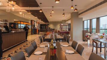 Un restaurante moderno con una larga mesa de comedor lista para una comida, una zona de bar y vistas de la ciudad a través de amplios ventanales.