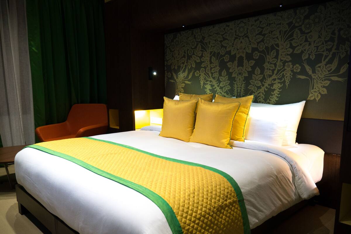 Una habitació d'hotel amb un llit gran, un llençol de llit verd i groc, un reposacapses de fusta i una cortina verda.