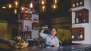 Zona de bar con un cantinero sirviendo una bebida, un frutero y estantes llenos de botellas de licor.
