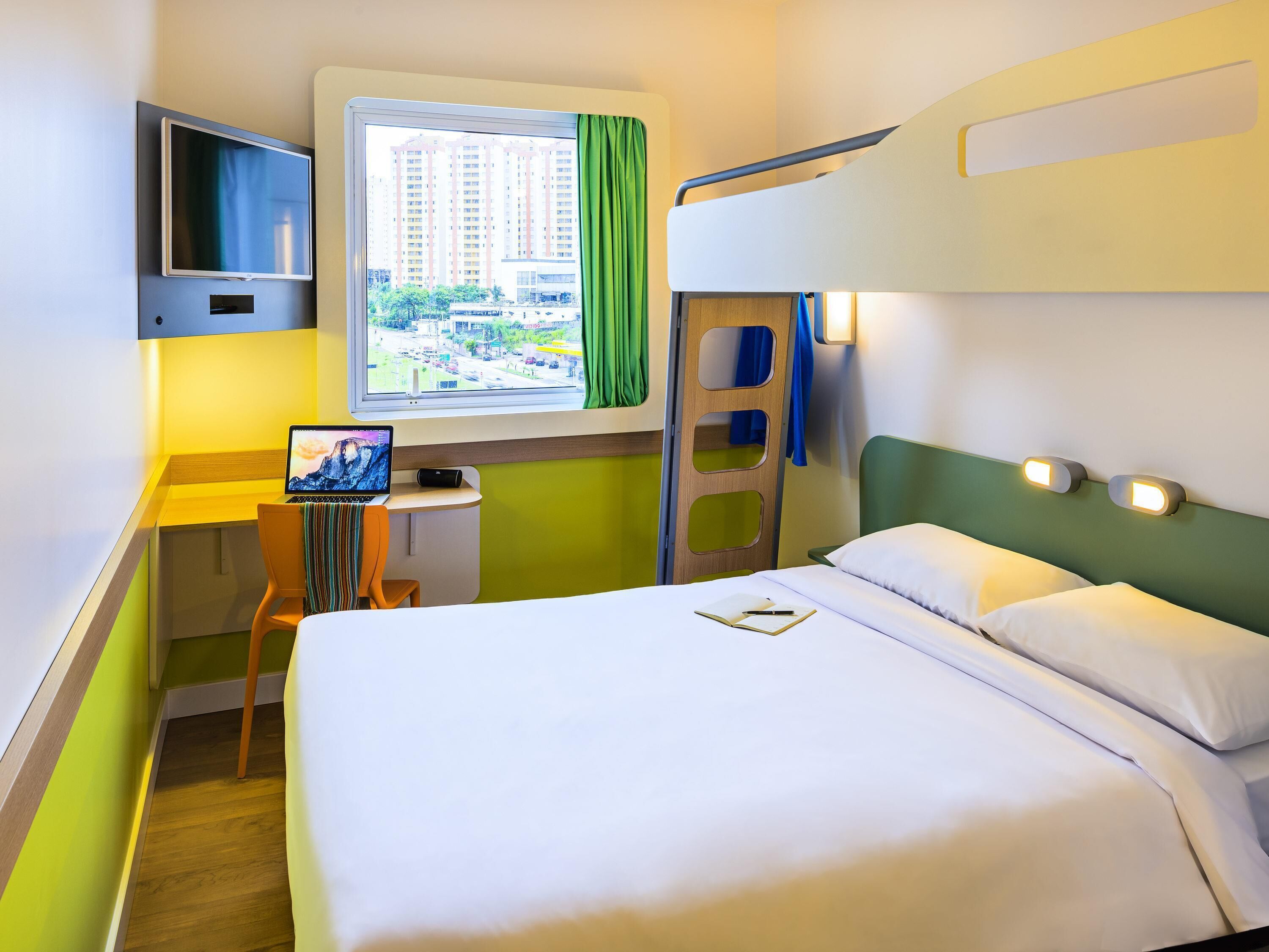 A hotel room with a bed, a desk with a laptop, a TV, and a window with a view of buildings.