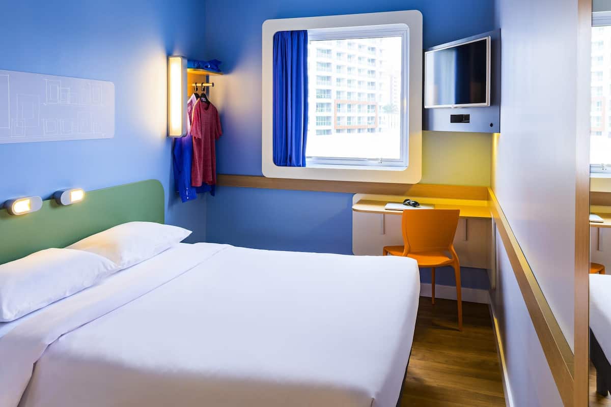 Habitación de hotel con cama, escritorio, silla, televisor y ventana con vista a edificios.