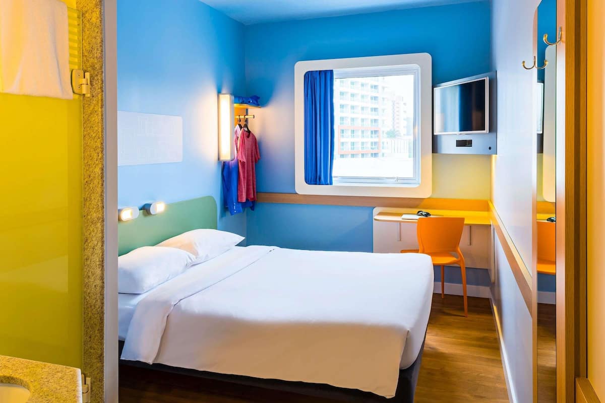 A hotel room with a bed, a desk, a chair, a TV, and a window with a curtain.