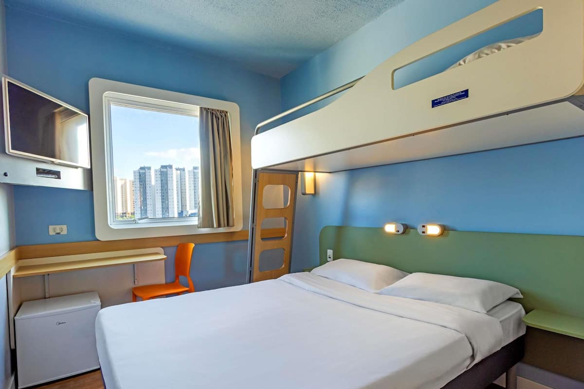 Habitación de hotel con literas, un escritorio con silla, un refrigerador pequeño y ventana con vista a edificios.