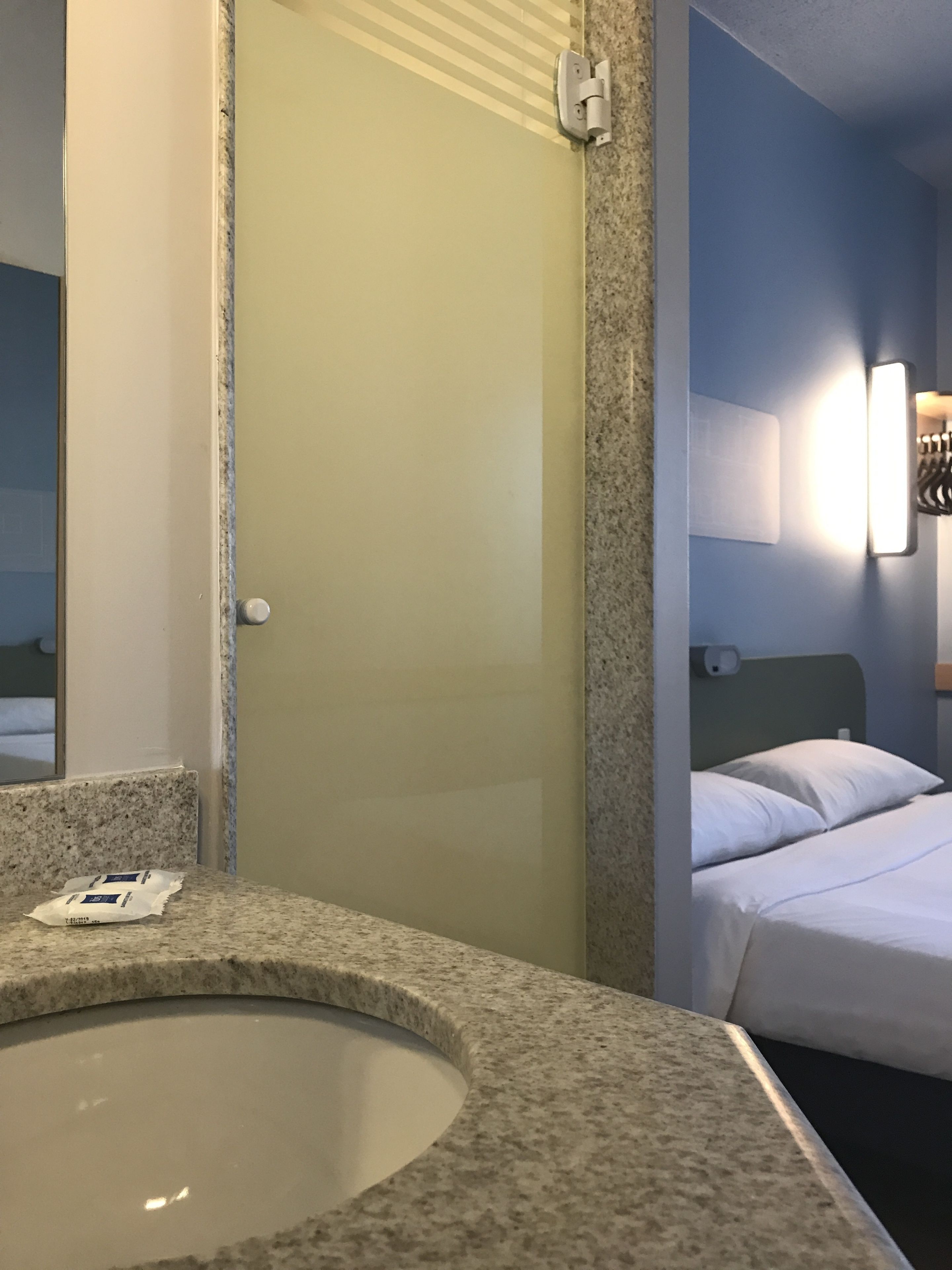 A hotel room with a granite countertop, a sink, a bed with white bedding, and a wall-mounted light fixture.
