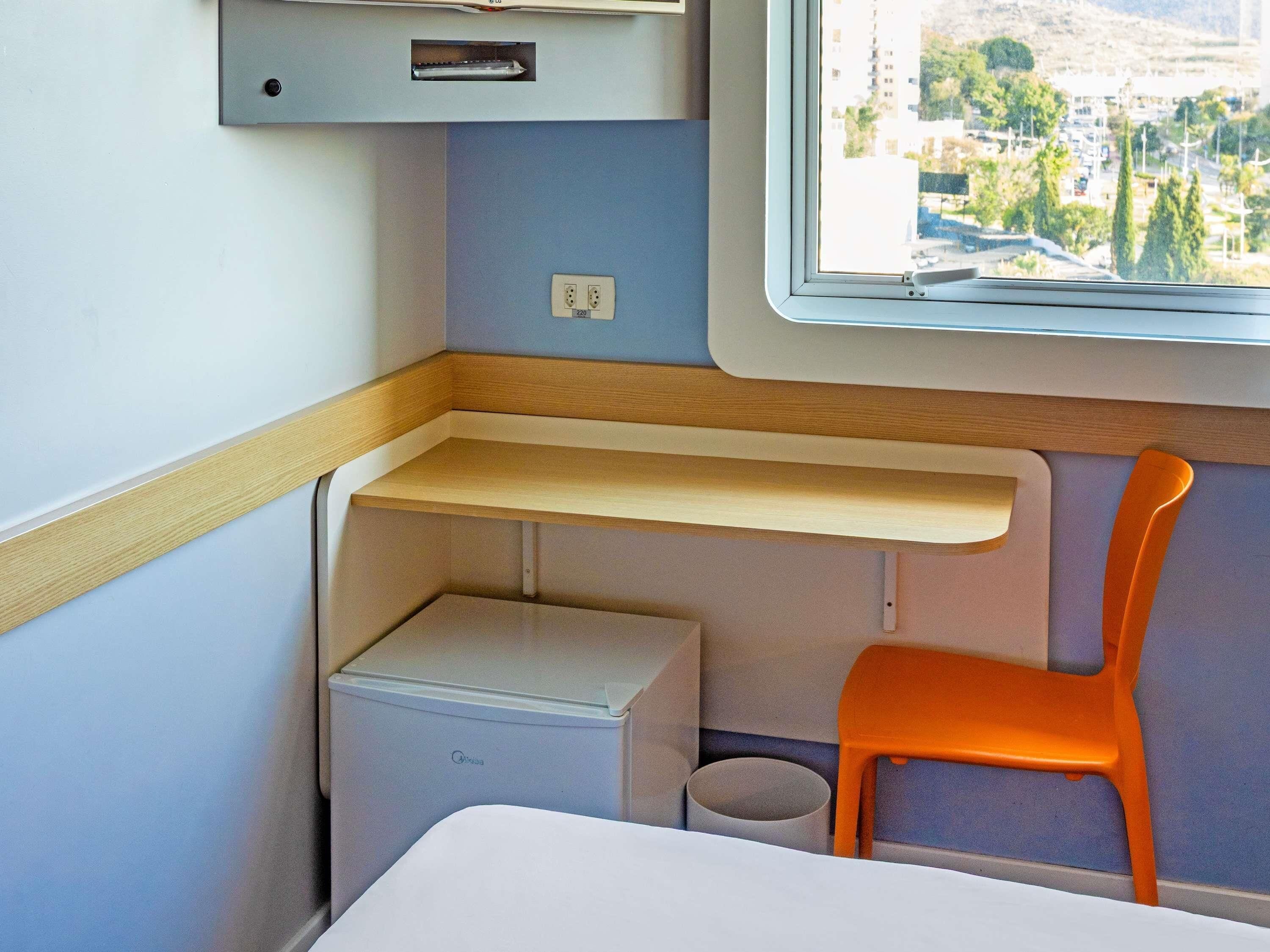 Um compartimento de trem compacto com uma pequena mesa, uma cadeira, uma prateleira, uma caixa de armazenamento e uma janela com vista para a cidade.