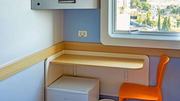 Um compartimento de trem compacto com uma pequena mesa, uma cadeira, uma prateleira, uma caixa de armazenamento e uma janela com vista para a cidade.
