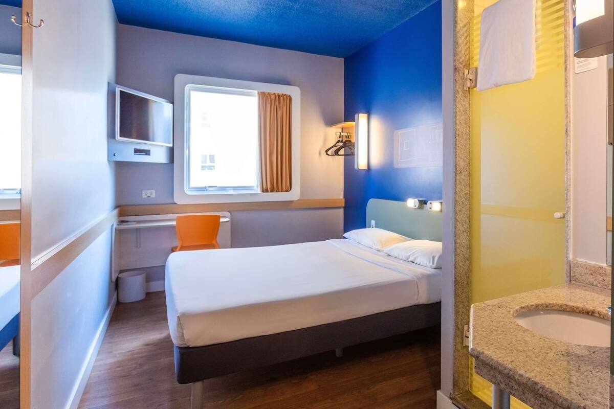 Habitación de hotel con cama, escritorio, silla, ventana con cortinas, televisor y baño con lavamanos.
