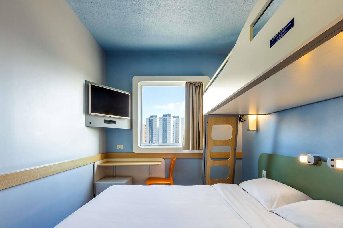 Habitación de hotel con cama, escritorio, silla, televisor, ventana y puerta, y vista a la ciudad.