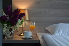 A neatly made bed with white linens, a bedside table with a lamp, a cup of coffee, a glass of orange juice, and a vase with purple flowers.
