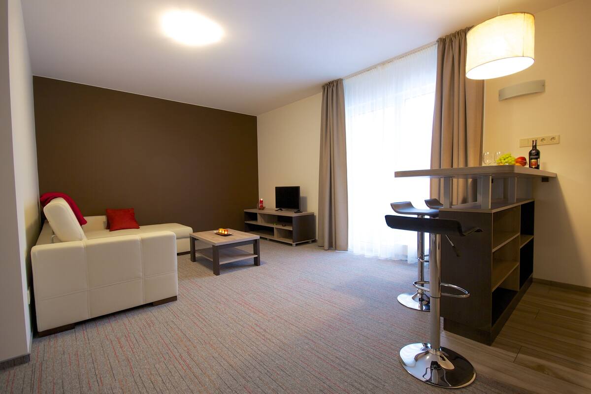 A modern living room with a white sofa, a small coffee table, a TV, and a bar area with a stool.