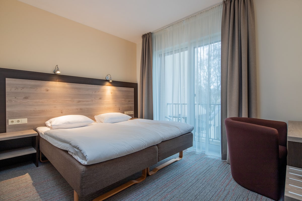 A hotel room with a bed, bedside tables, a chair, and a large window with curtains.