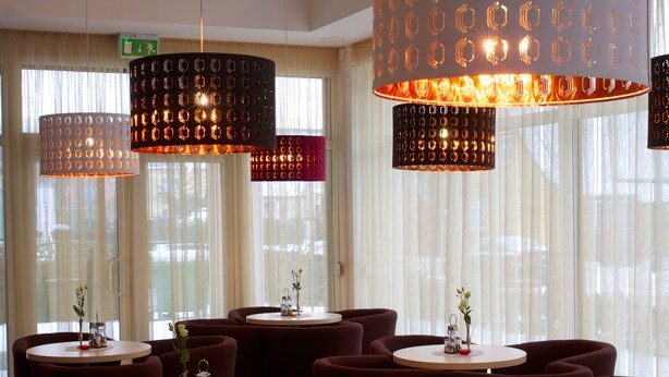 Ein Restaurant mit runden Tischen und Stühlen, großen hängenden Pendelleuchten und Blick durch transparente Vorhänge.