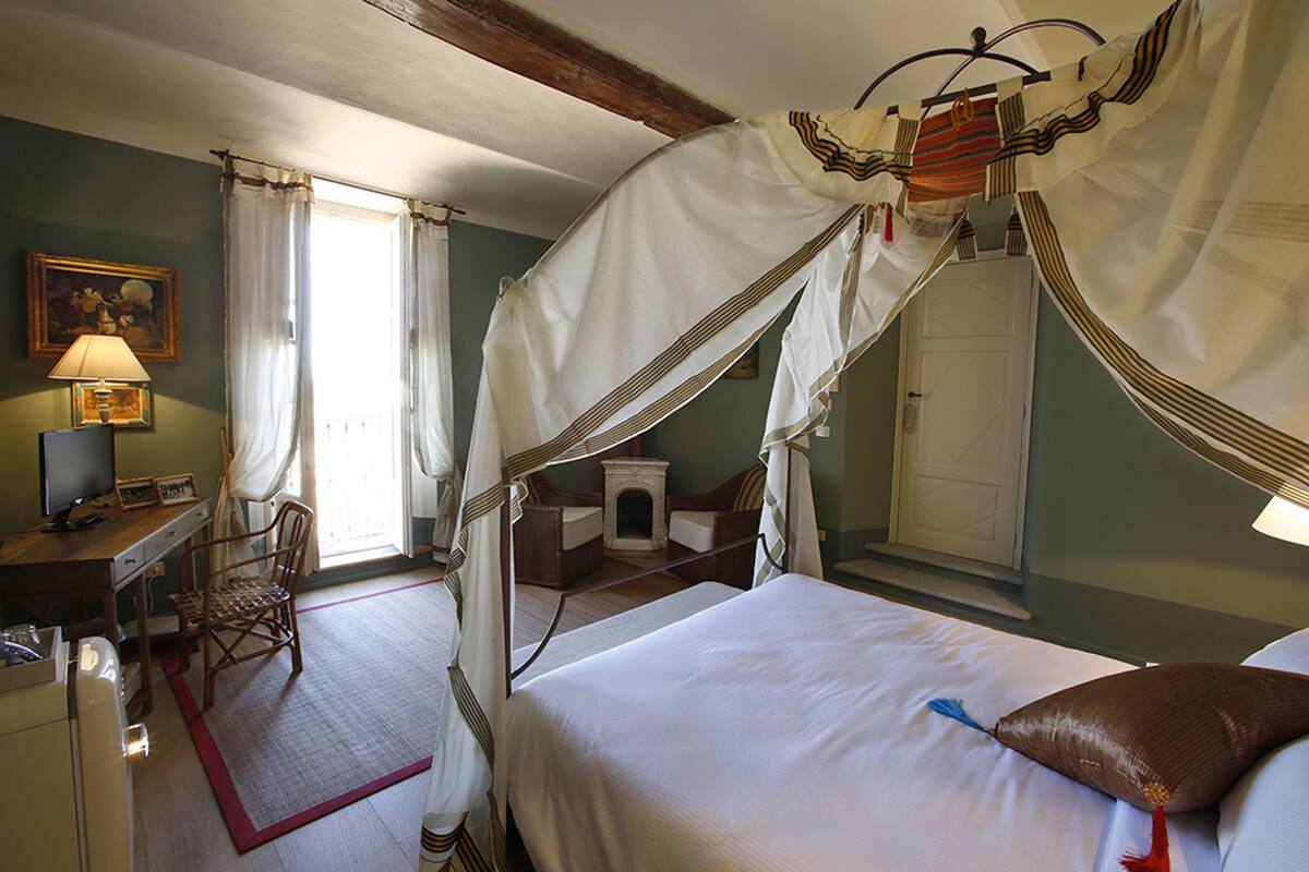 Een kamer met een hemelbed, een bureau met een lamp, een stoel en een kleine open haard.