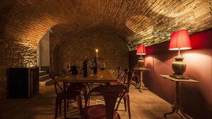 Une pièce faiblement éclairée, avec un plafond en briques, une longue table dressée pour le repas et deux lampes dotées d’abat-jour rouges.