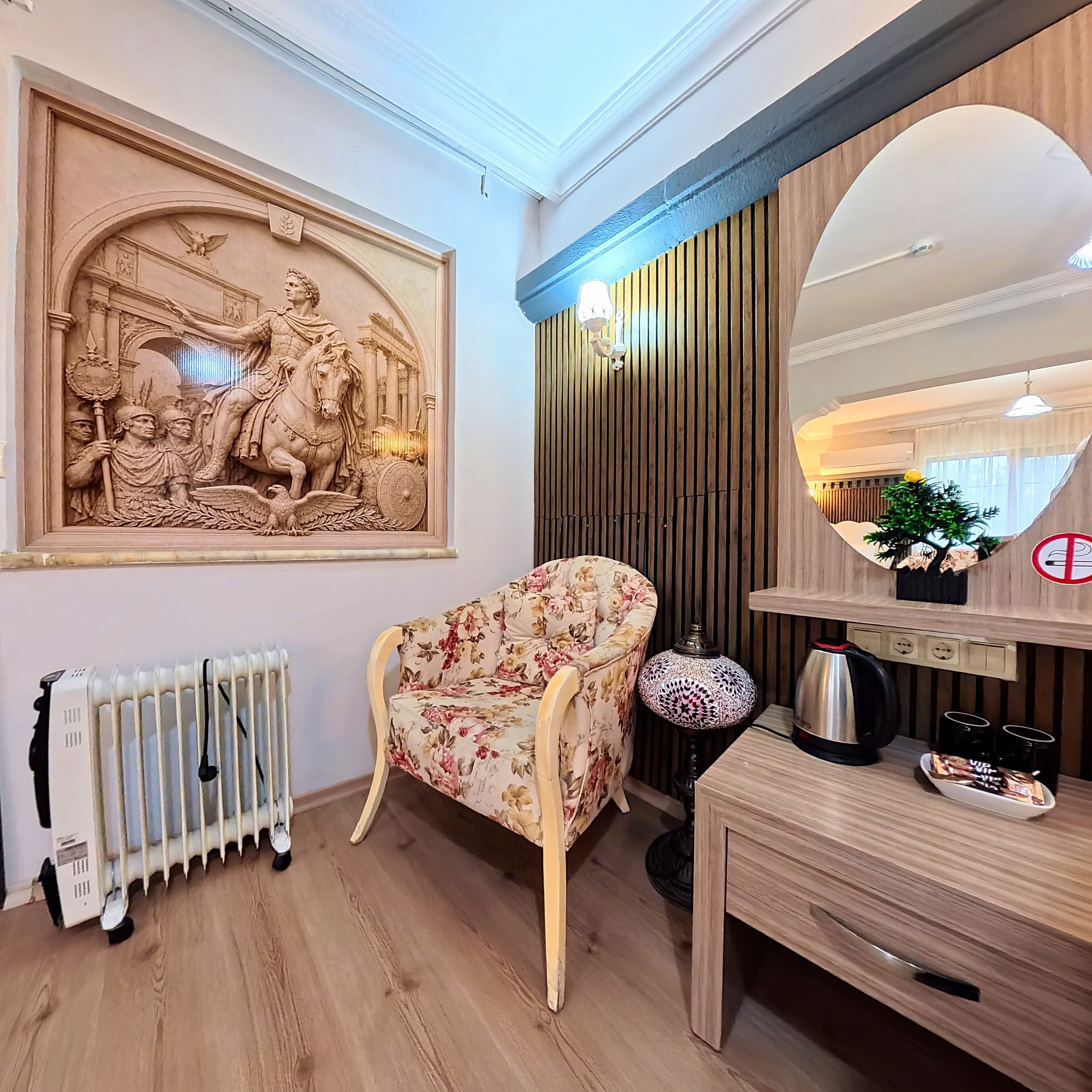 Una habitación con un radiador, un sillón estampado con flores, un escritorio de madera con un espejo y una escultura en la pared.