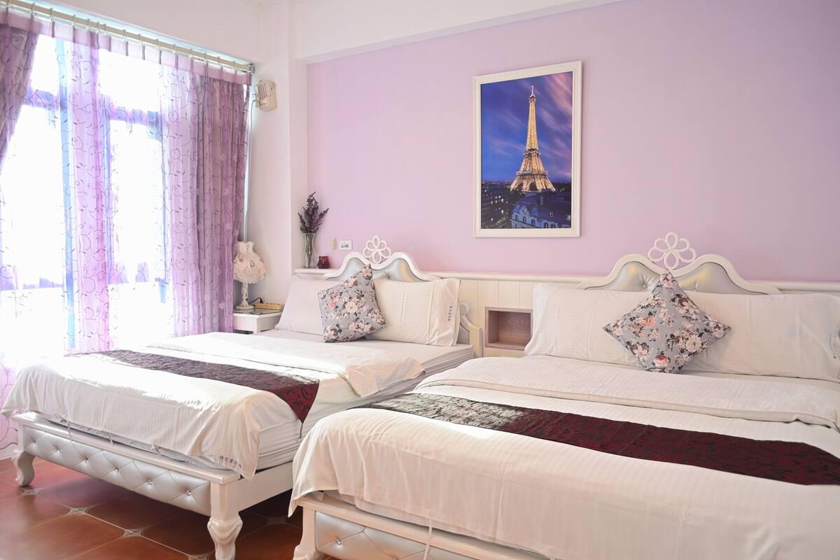 Habitación de hotel con dos camas, un ventanal grande con cortinas, un cuadro colgado en la pared de la Torre Eiffel y una mesita de noche con un florero.