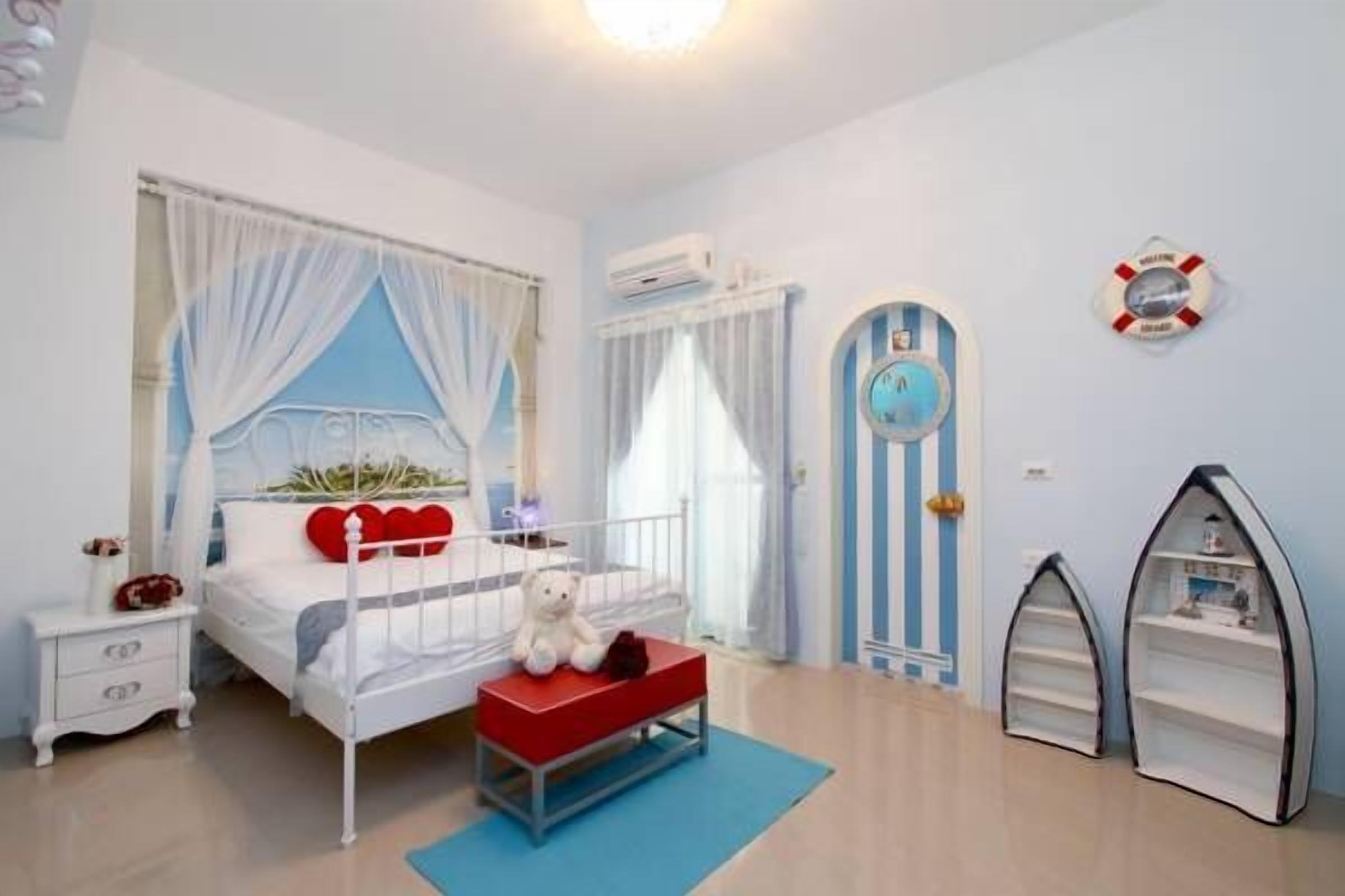 A bedroom with a bed, bedside table, and a red bench. There is a window with curtains, a wall clock, and a life preserver decoration.
