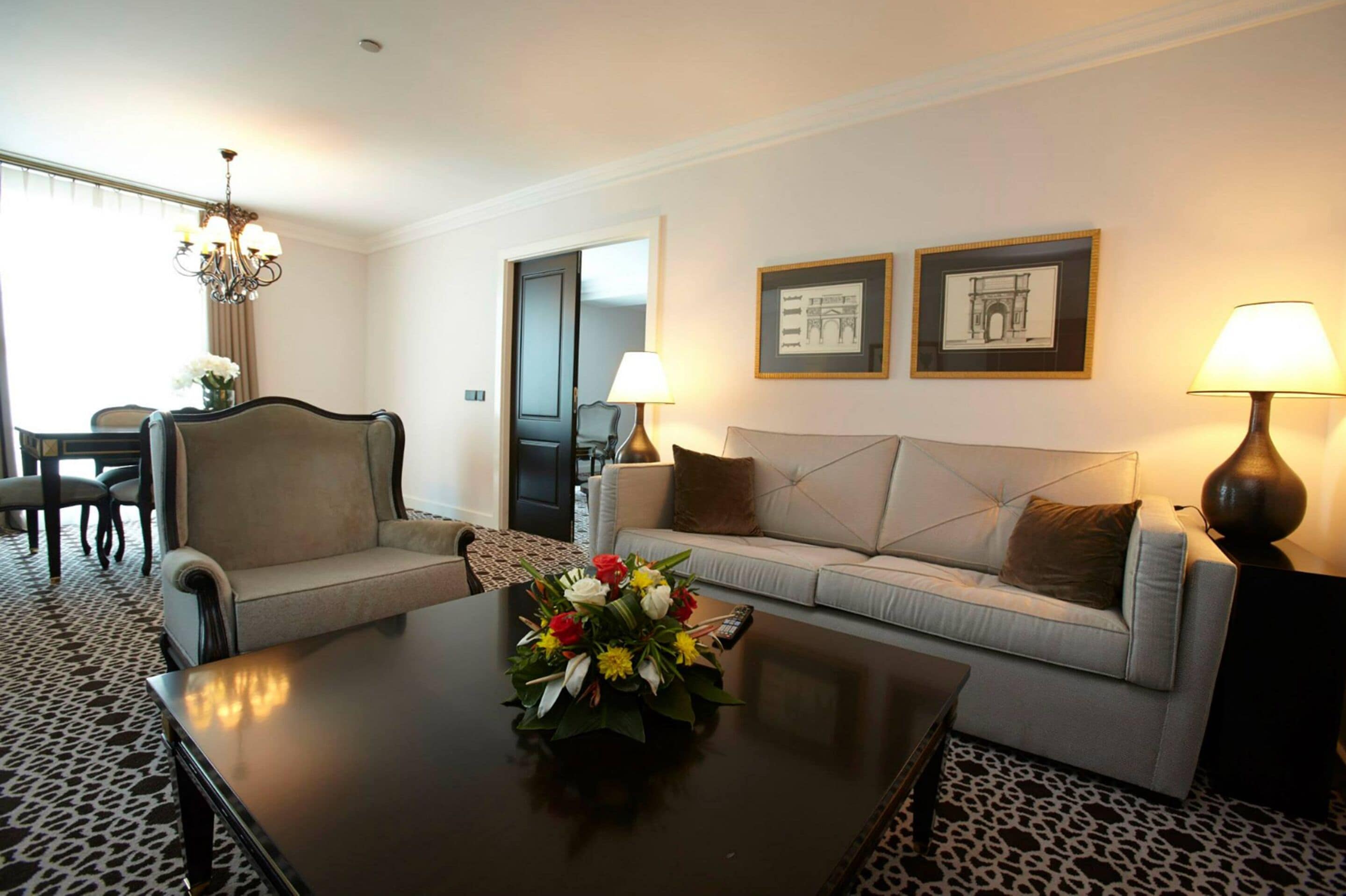 A living room with a sofa, armchair, coffee table, and two lamps. There are two framed pictures on the wall and a chandelier.