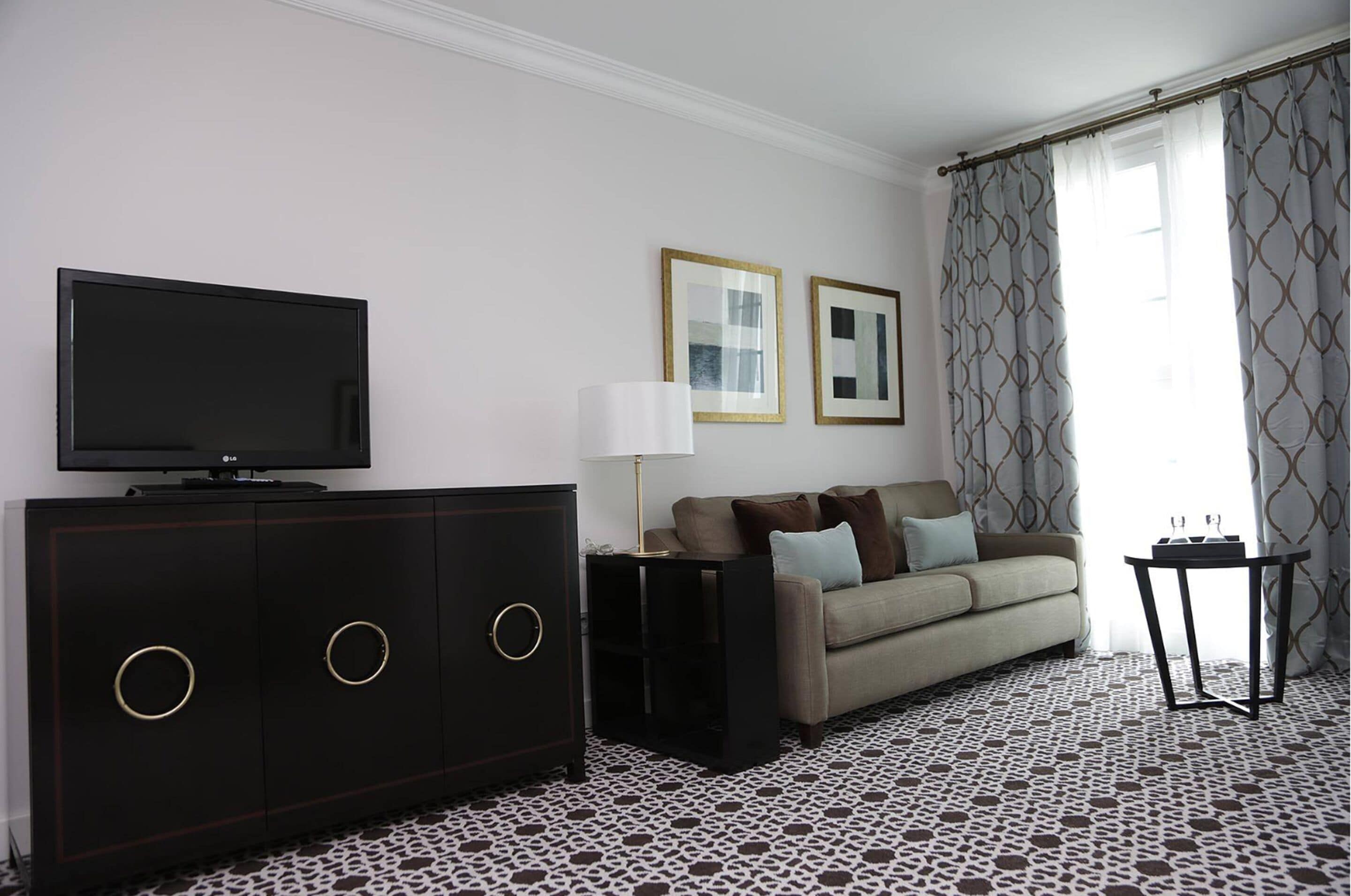 Un salón con televisor de pantalla plana, un mueble de madera oscura con pomos redondos, un sofá beige con cojines, una mesita con una lámpara y cortinas estampadas.