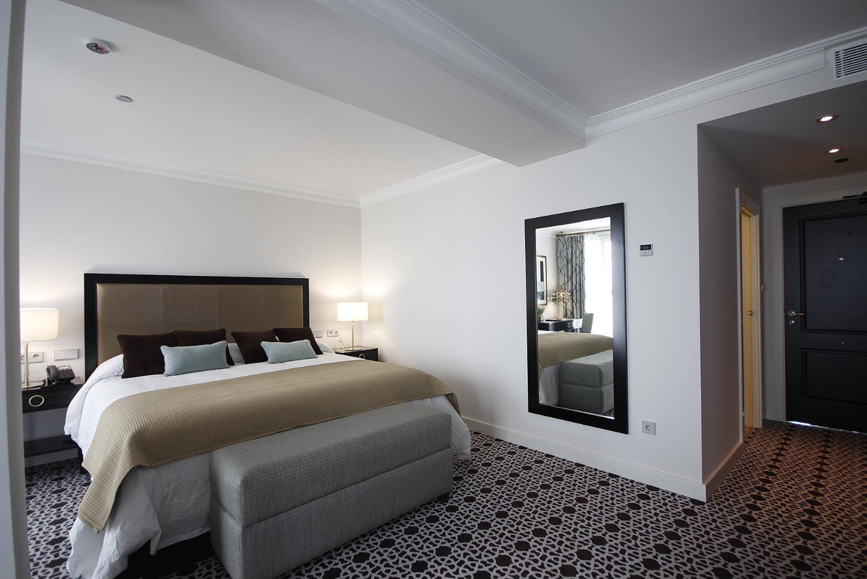 Quarto de hotel com uma cama grande, mesas de cabeceira, um espelho e um chão com padrão.