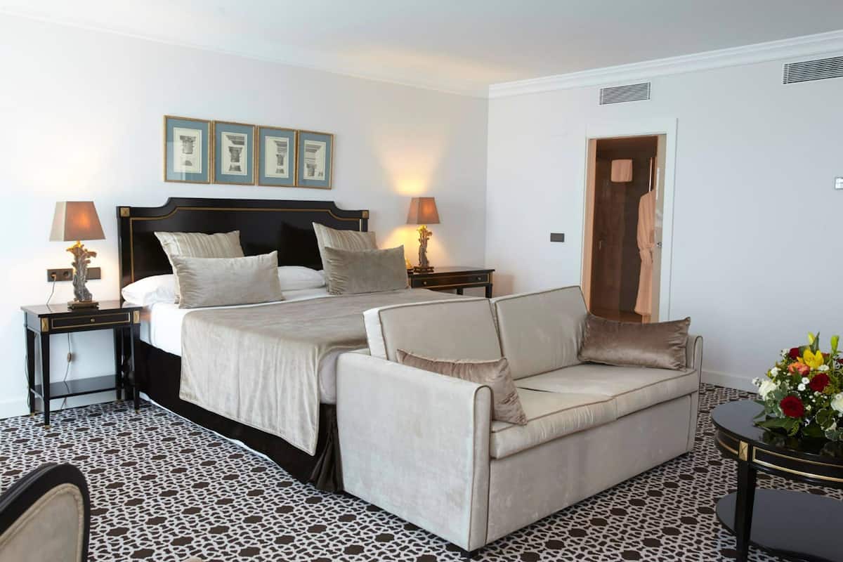 Habitación de hotel con cama doble, dos mesitas de noche con lámparas, un sofá, piso con diseño y cuadros en la pared.