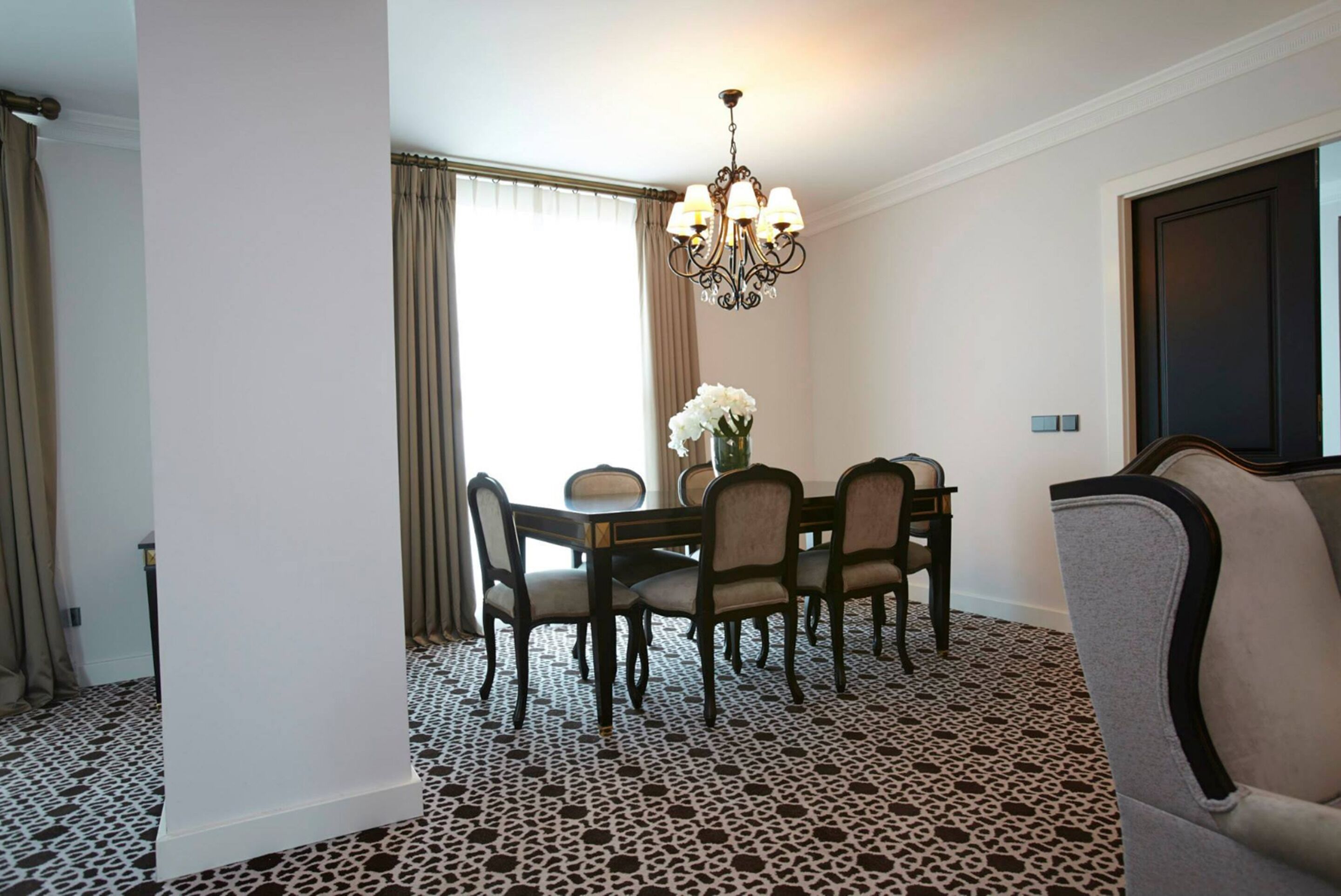A dining area with a round table, six chairs, a chandelier, and a vase with white flowers.