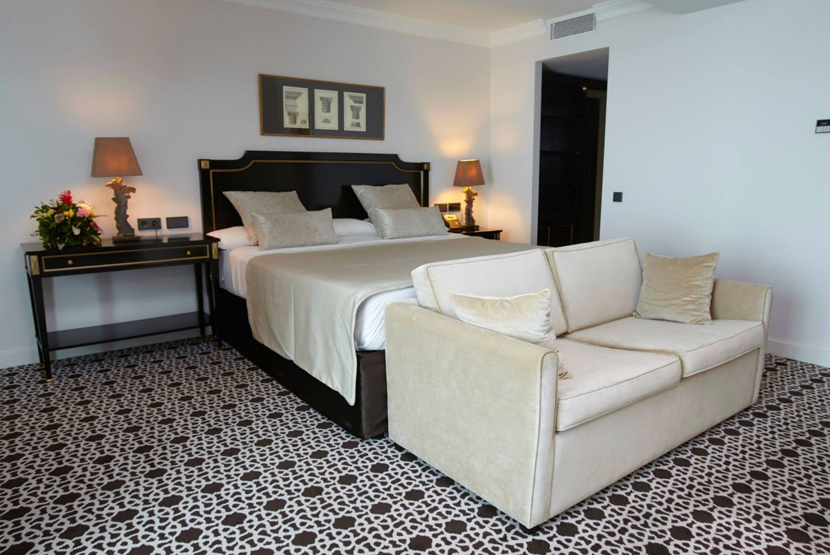 A hotel room with a bed, a sofa, a nightstand with lamps, and a patterned floor.