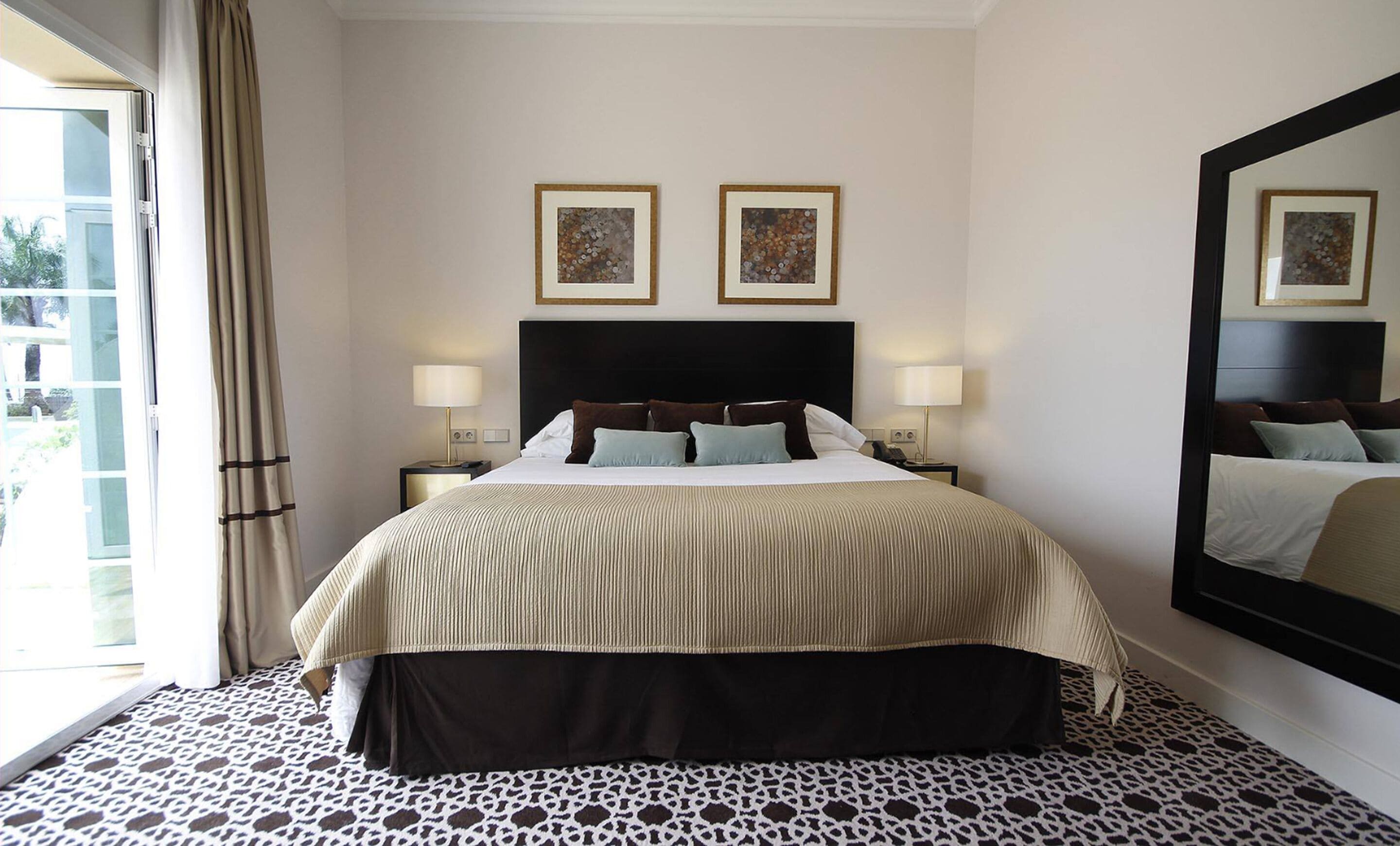 A hotel room with a bed, two bedside lamps, a large mirror, and two framed pictures on the wall.
