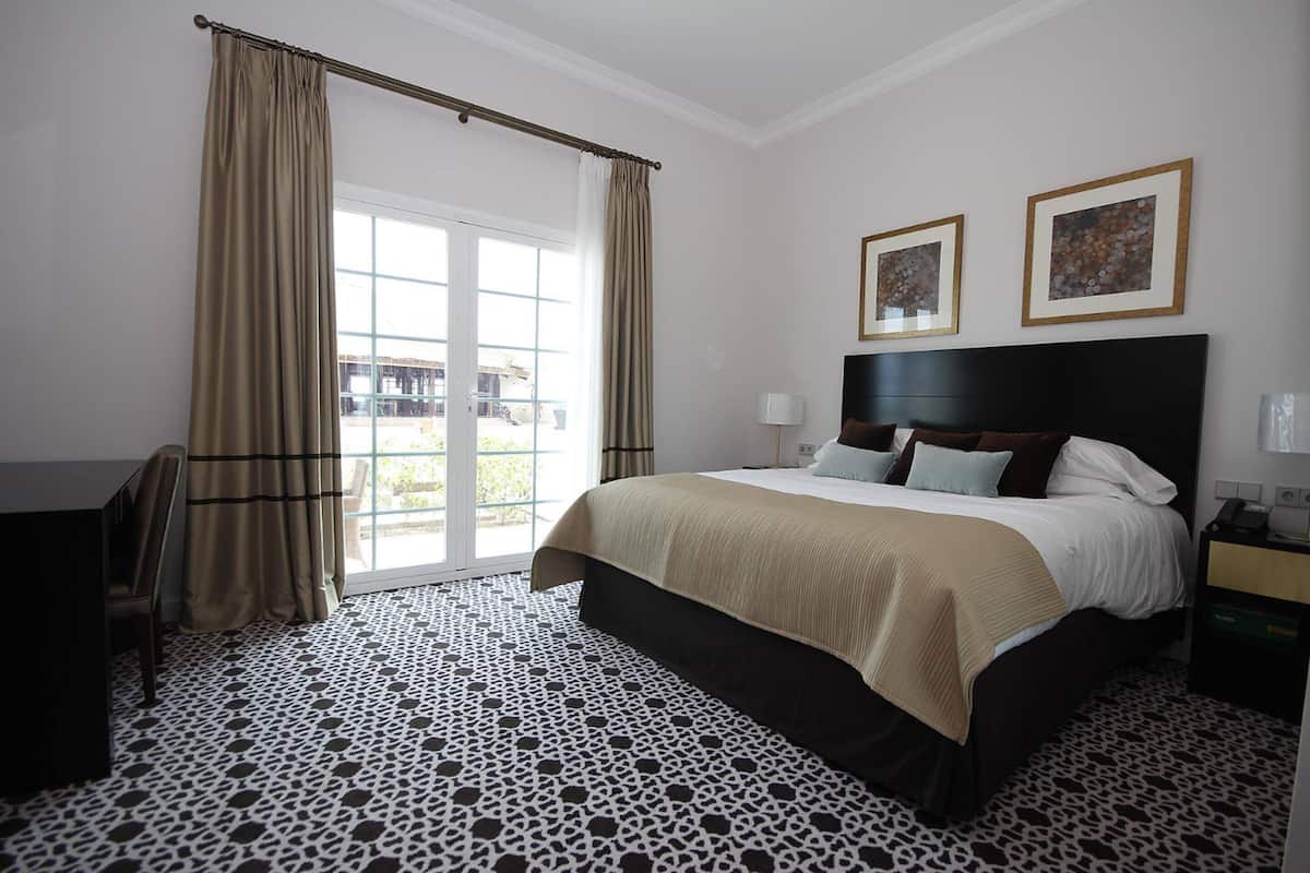 Habitación de hotel con una cama grande, un escritorio, una silla, un ventanal con cortinas y dos cuadros en la pared.