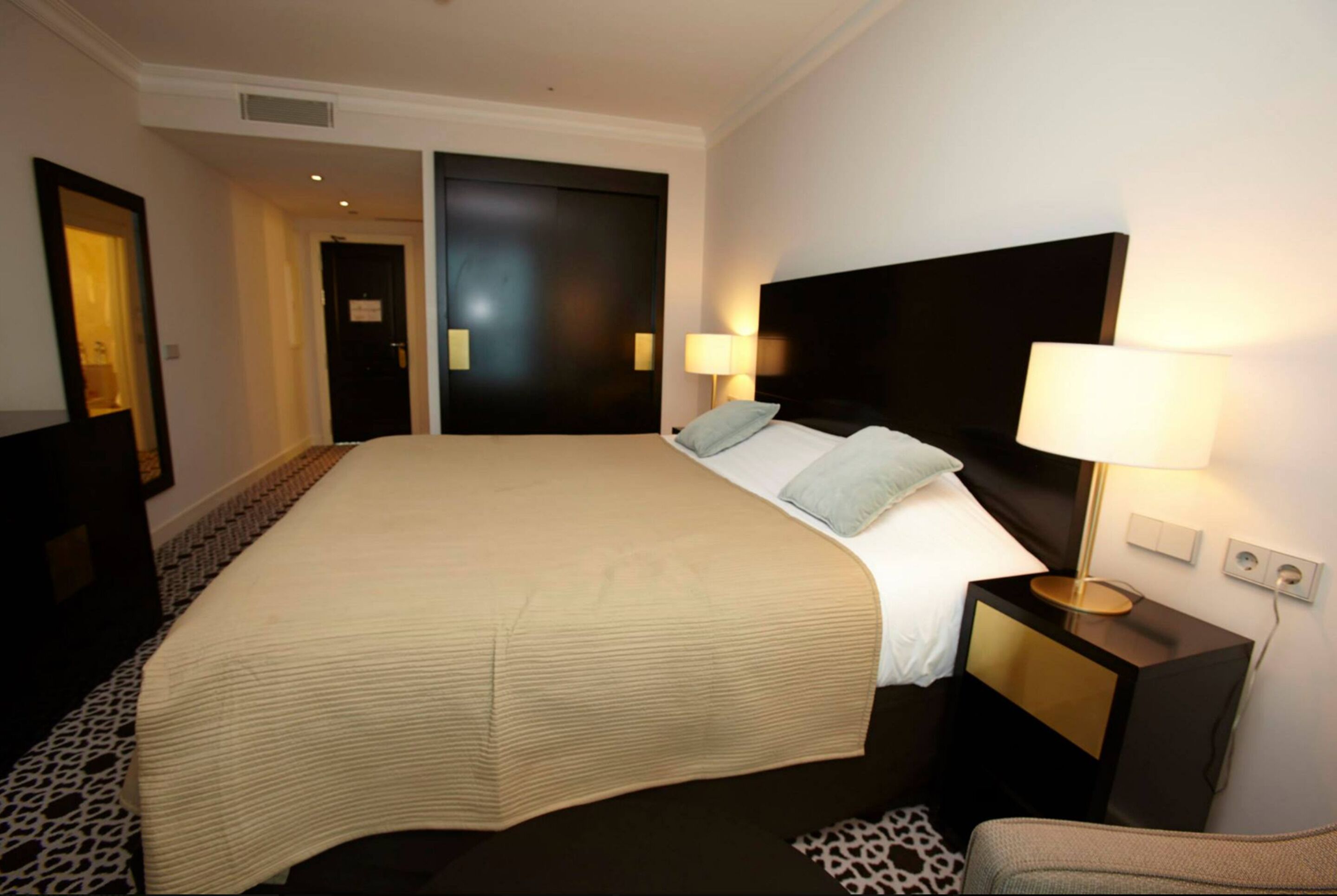Una habitación de hotel con una cama grande, dos lámparas de noche, una mesita de noche y una alfombra estampada.